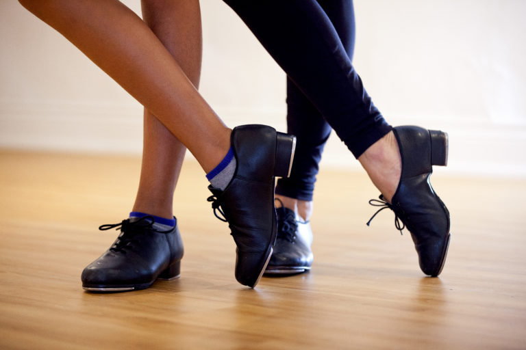 从脚踝以下，两个穿着黑色踢踏舞鞋的舞者一只脚交叉在另一只脚上