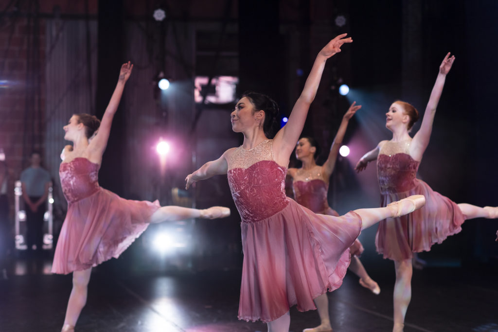 四名芭蕾舞演员穿着玫瑰色的当代芭蕾舞裙，摆着piqué阿拉伯式的低姿势。