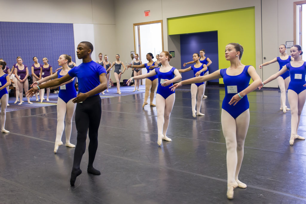 一群芭蕾舞演员正在表演“腾空而起”。他们穿着蓝色的紧身衣。在这群人的前面是一个黑人男舞者。