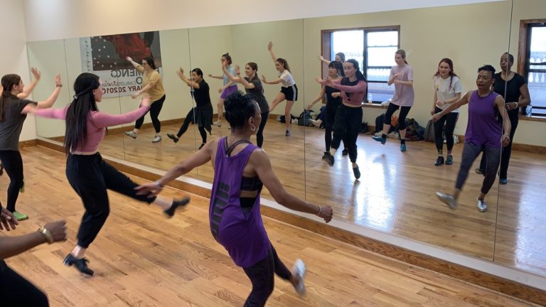 杰曼·古德森，一个穿着紫色背心的黑人妇女，教一个踢踏舞班。她和学生们把一条腿伸到前面。我们从后面看到他们，看到他们在镜子里的倒影。