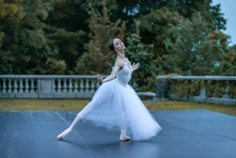 一个穿着白色长芭蕾舞裙的芭蕾舞女演员在户外的舞台上跳舞。