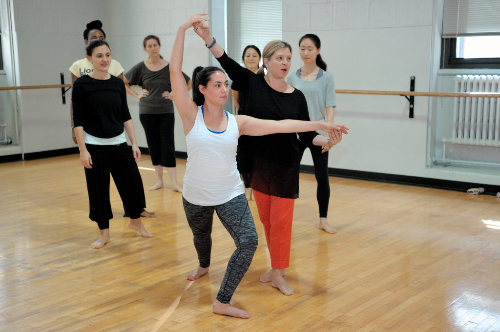 黛博拉·达马斯特身穿黑色长袖上衣和橙色裤子，正在指导一名舞蹈研究生，背景是其他五名学生。